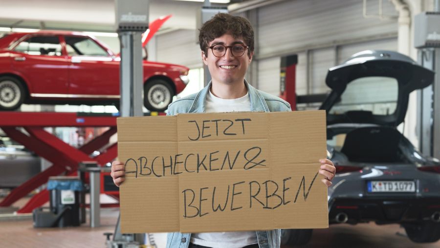 Ein lächelnder Mann steht in einer Werkstatt und einem Schild in beiden Händen mit dem Text: Jetzt abchecken & bewerben