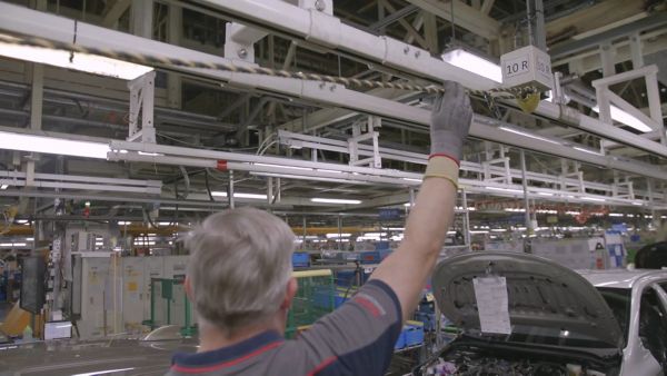 Person mit Handschuhen steht in einer Fabrikhalle und hält die Hand an einer Schnur über ihrem Kopf