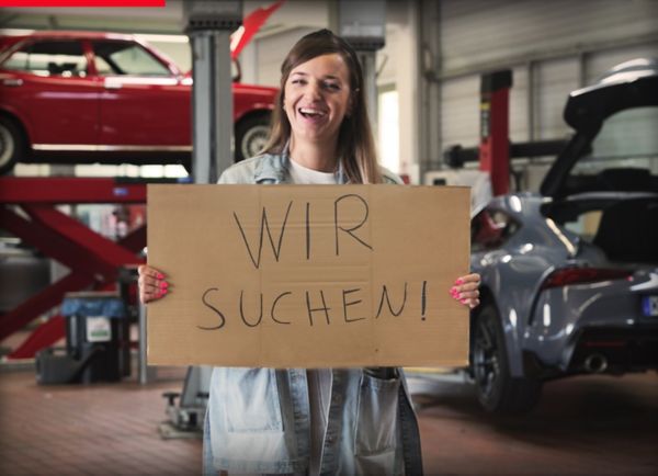 Eine Person in einer Werkstatt hält ein Schild in die Kamera. Darauf steht: Wir suchen!