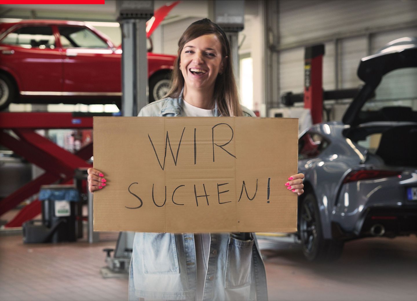 Eine lächelnde Frau steht in einer Werkstatt und hält ein Schild in der Hand mit dem Text: Wir suchen!