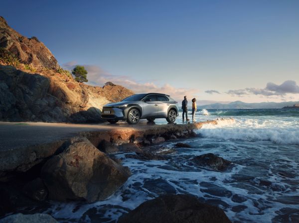 Ein silberner Toyota bZ4X steht an einer welligen Küste, daneben stehen zwei Personen und schauen auf das Meer