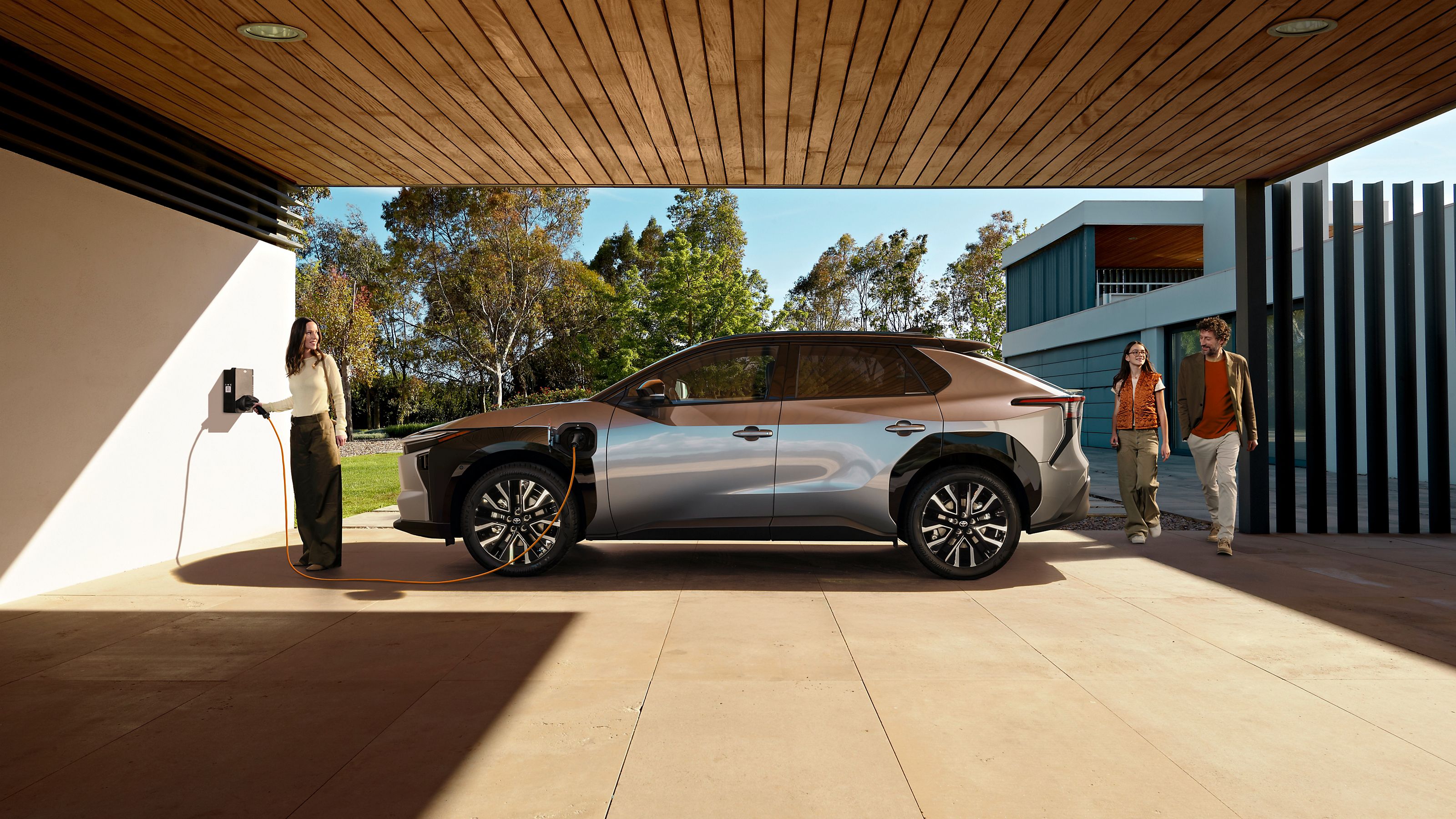 Eine Familie lädt ihren Toyota bZ4X über eine Wallbox, die unter einem Carport mit Holzdecke vor einem modernen Neubau steht.