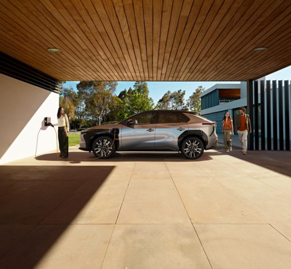 Eine Familie lädt ihren Toyota bZ4X über eine Wallbox, die unter einem Carport mit Holzdecke vor einem modernen Neubau steht.