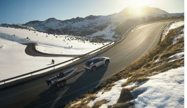 Ein silber-schwarzer Toyota bZ4X zieht einen Anhänger mit einem weißen Schlauchboot eine kurvige Landstraße hoch, während im Hintergrund ein großer Berg zu sehen ist, an dessen Spitze die Sonne untergeht.