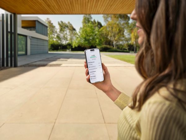 Eine Frau, die in einem überdachten Carport vor einem modernen Neubau steht, schaut auf ihrem Handy den Batteriestatus mit der MyToyota-Smartphone-App an.