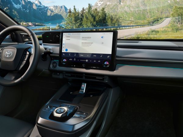 Blick in das Innere des neuen Toyota bZ4X, der einen Blick auf das elegante, dunkle Cockpit mit sehr großem Display zeigt, während das Auto auf einer bergigen Landstraße durch die Landschaft fährt.