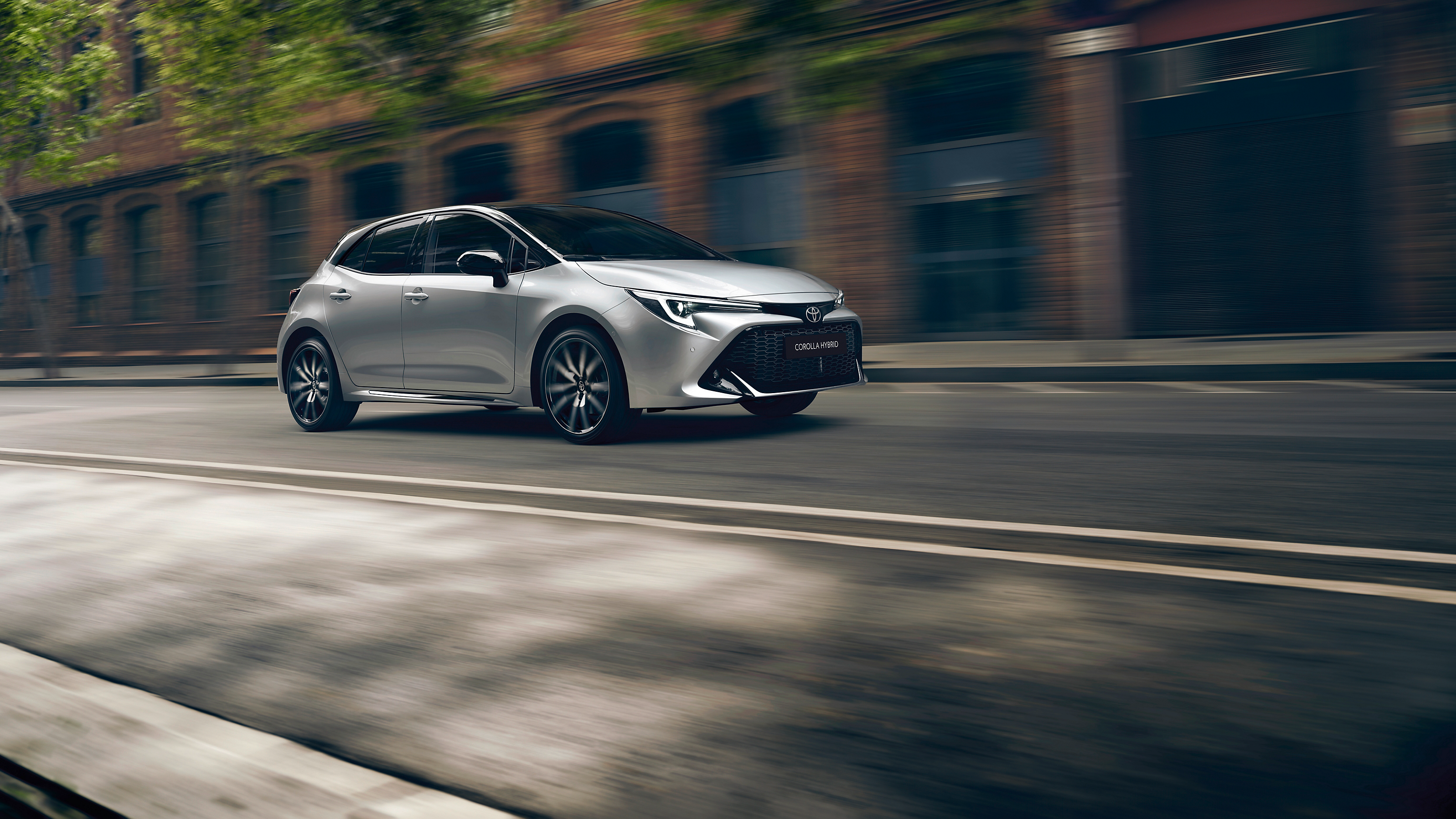 A Toyota Corolla Hatchback drives along a tree-lined city street