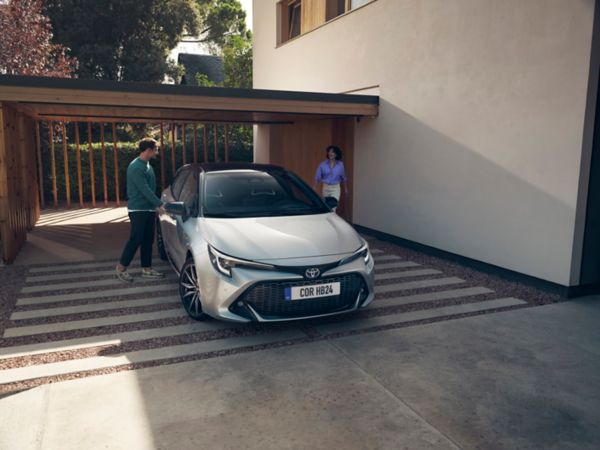 Zwei Personen steigen in einen silbernen Corolla ein, der auf unter einem Carport neben einem Haus geparkt ist