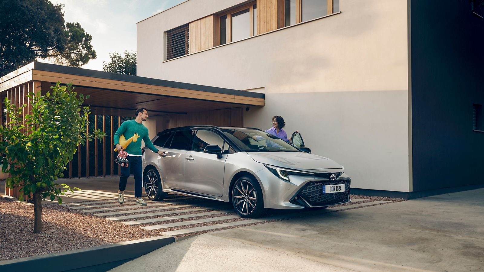 Zwei Personen steigen in einen silbernen Corolla Touring Sports, der auf einer Auffahrt unter einem Carport steht