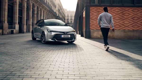 Ein silberner Toyota Corolla von 2024 lässt blinkend vor dem Abbiegen einen Mann passieren, im Hintergrund Gebäude mit Backstein und Rundbögen