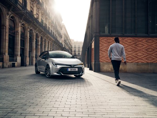 Person läuft einen silbernen Corolla zu, der auf einem gepflasterten Platz zwischen zwei Gebäuden geparkt ist