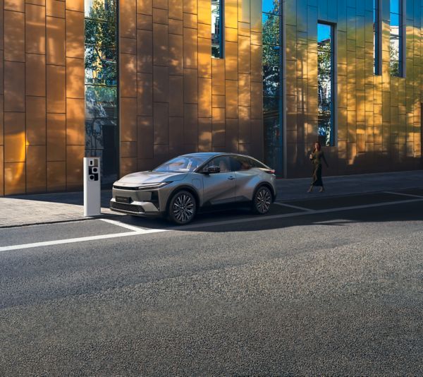 Ein Toyota C-HR+ wird an einer öffentlichen Ladestation auf der Straße vor einem reflektierenden Gebäude aufgeladen.