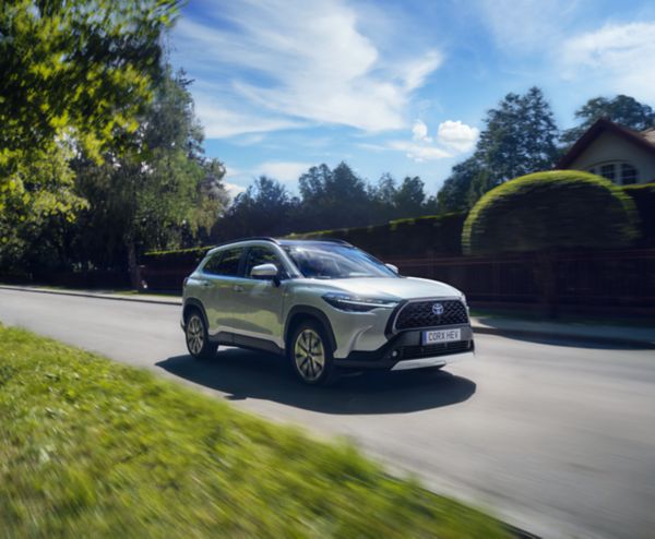 Silberner Toyota Corolla Cross fährt auf einer Landstraße und wird von der Sonne angestrahlt