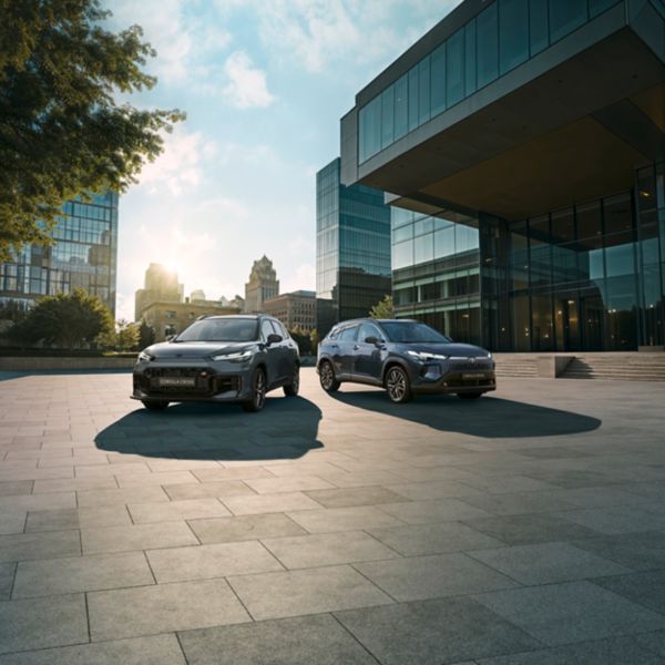 Vor einem Bürokomplex parken zwei Corolla Cross, wie sie häufig als Firmenautos genutzt werden, wenn Mitarbeiter zu Terminen oder Kundengesprächen unterwegs sind.