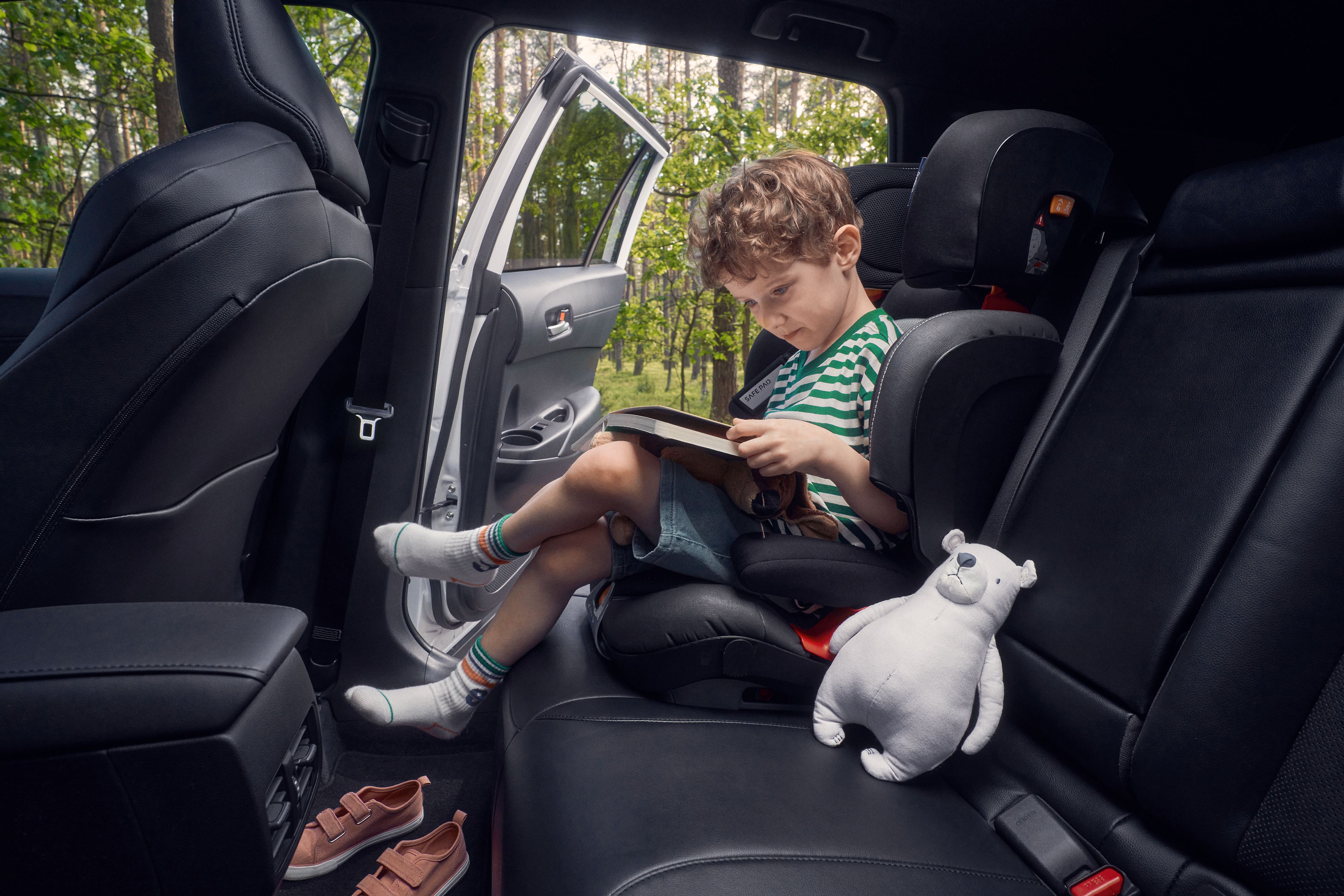Kind sitzt mit überschlagenen Beinen auf der Rückbank auf einem Kindersitz und schaut auf ein Buch