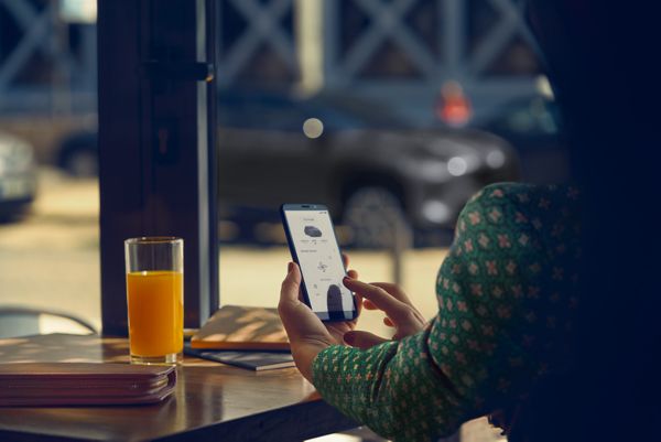 Lady sat at a restaurant table using the TOYOTA MyT app on her phone, her Toyota Yaris Cross is parked in the background.