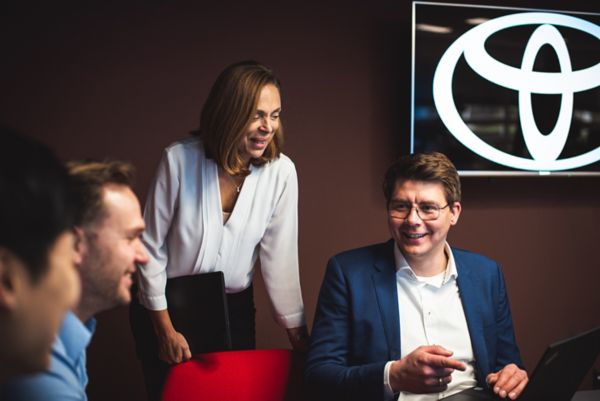 Personen sitzen in einem Meeting und lächeln. Im Hintergrund ist ein Fernseher mit Toyota Logo zu sehen. 