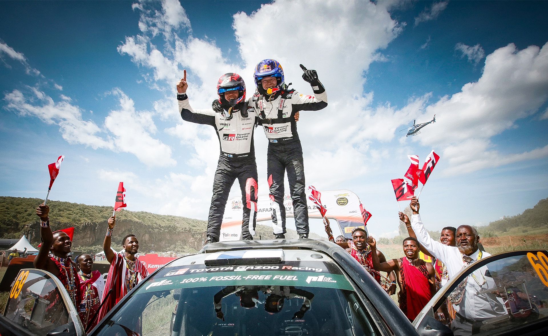 Dwaj kierowcy rajdowi stoją na dachu Toyoty GR i cieszą się ze zwycięstwa wśród kibiców.