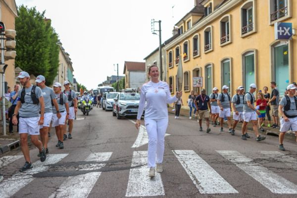 Carina Hoffmann, gekleidet in weiß, führt eine Gruppe Läufer mit einer Fackel über einen Zebrastreifen, im Hintergrund sind Fahrzeuge und Zuschauer zu sehen.