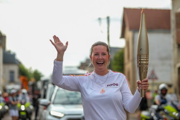 Carina Hoffmann, weiß gekleidet, winkt lachend mit einer goldenen Fackel in der Hand, im Hintergrund unscharf Begleitfahrzeuge und Motorräder