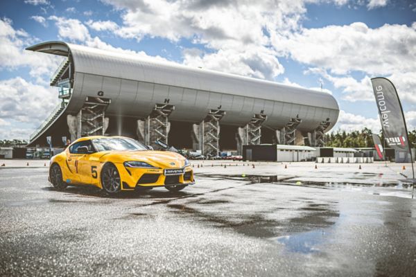 Ein gelber Sportwagen driftet auf einer nassen Strecke, im Hintergrund ist eine Tribüne neben einer Rennstrecke zu sehen