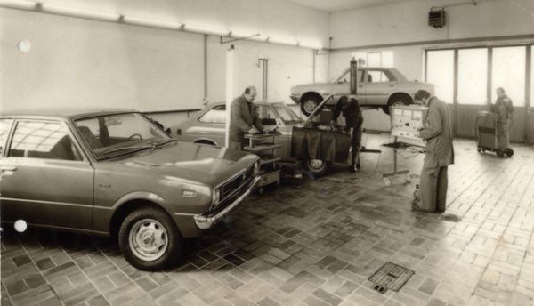 Schwarzweiß-Bild von der Werkstatt mit mehreren alten Toyota-Modellen und Mitarbeitern