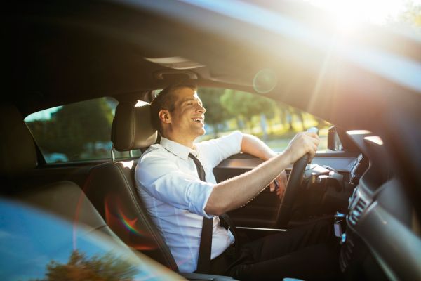 Ein Mann in weißem Hemd und Krawatte lächelt, während er ein Auto fährt, und Sonnenlicht strömt durch die Fenster.