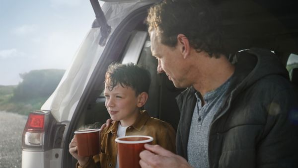 Zwei Personen, ein Kind und ein Erwachener, sitzen im geöffneten Kofferraum eines großen Autos und trinken eine heiße Tasse Tee