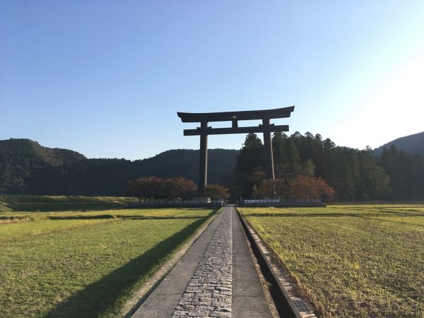 Ein großes traditionelles japanisches Tor steht am Ende eines steinernen Weges, umrahmt von üppigen grünen Feldern und fernen Bergen unter einem klaren blauen Himmel.