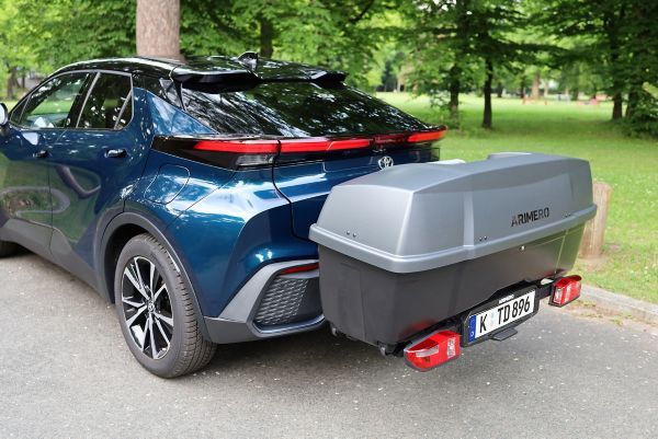 Blaues Fahrzeug von hinten mit einer grauen Heckbox, welches vor einer grünen Wiese mit Bäumen geparkt ist