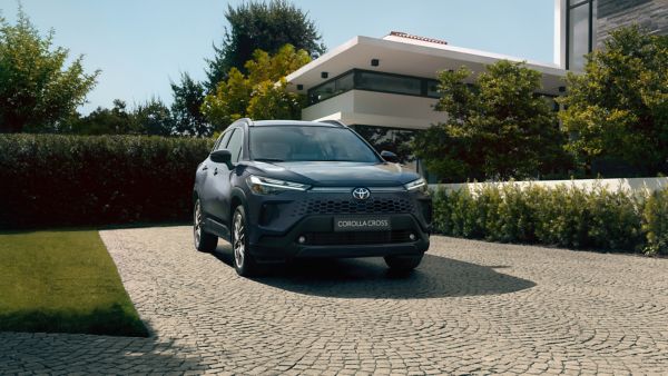 Widok z przodu ciemnoniebieskiego SUV-a Corolla Cross Hybrid zaparkowanego na kamiennym podjeździe przed dużym nowoczesnym domem otoczonym drzewami.