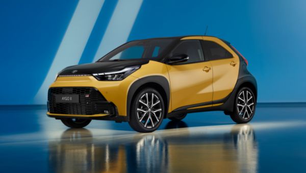 Egy Toyota Aygo X Hybrid kéttónusú metál arany színben, fekete motorháztetővel és tetővel, élénk kék stúdió környezetben.