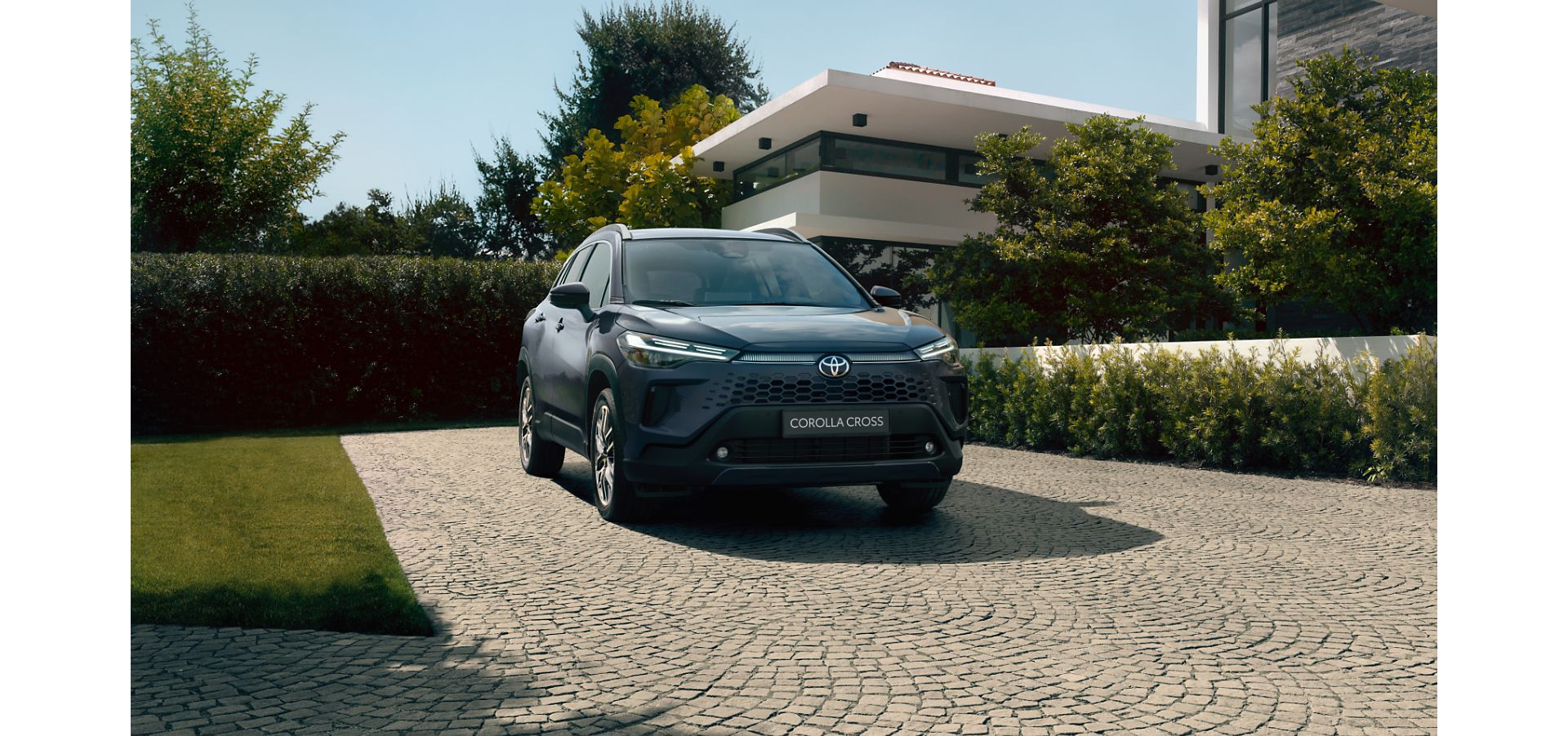 Front view of a dark blue Corolla Cross Hybrid SUV parked on a stone driveway in front of a large modern house framed by trees.