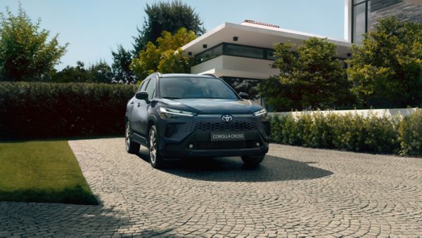 Widok z przodu ciemnoniebieskiego SUV-a Corolla Cross Hybrid zaparkowanego na kamiennym podjeździe przed dużym nowoczesnym domem otoczonym drzewami.