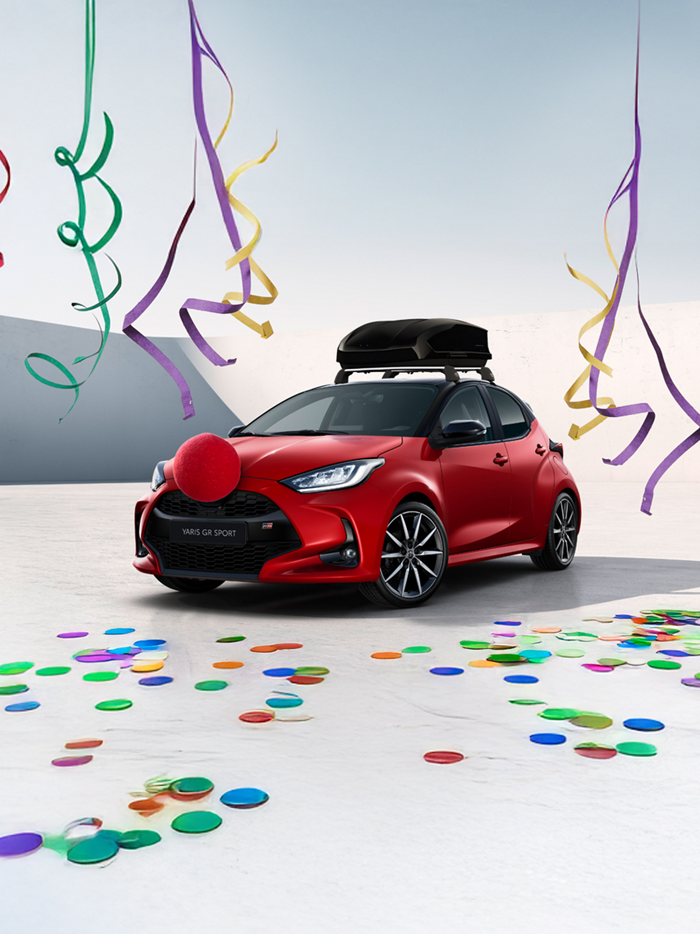 Toyota Yaris GR Sport in Rot mit schwarzer Dachbox und roter Clownsnase vor hellem Hintergrund, umgeben von bunten Luftschlangen und Konfetti auf dem Boden.