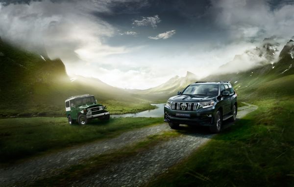 Zwei ältere Generationen des Land Cruisers fahren auf einer bergigen unbefestigen Strecke entlang. Im Hintergrund sieht man einen Bergkamm, der von Wolken bedeckt ist. 