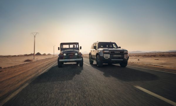 Toyota Landcruiser und altes Landcruiser Modell fahren nebeinander auf einer leeren Landstraße in einer Wüste.