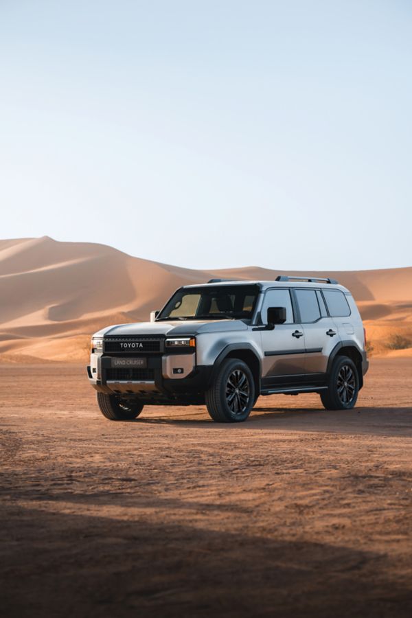 Land Cruiser steht in einer Wüste und wird von der Sonne angestrahlt. Im Hintergrund sieht man hohe Sanddünen.