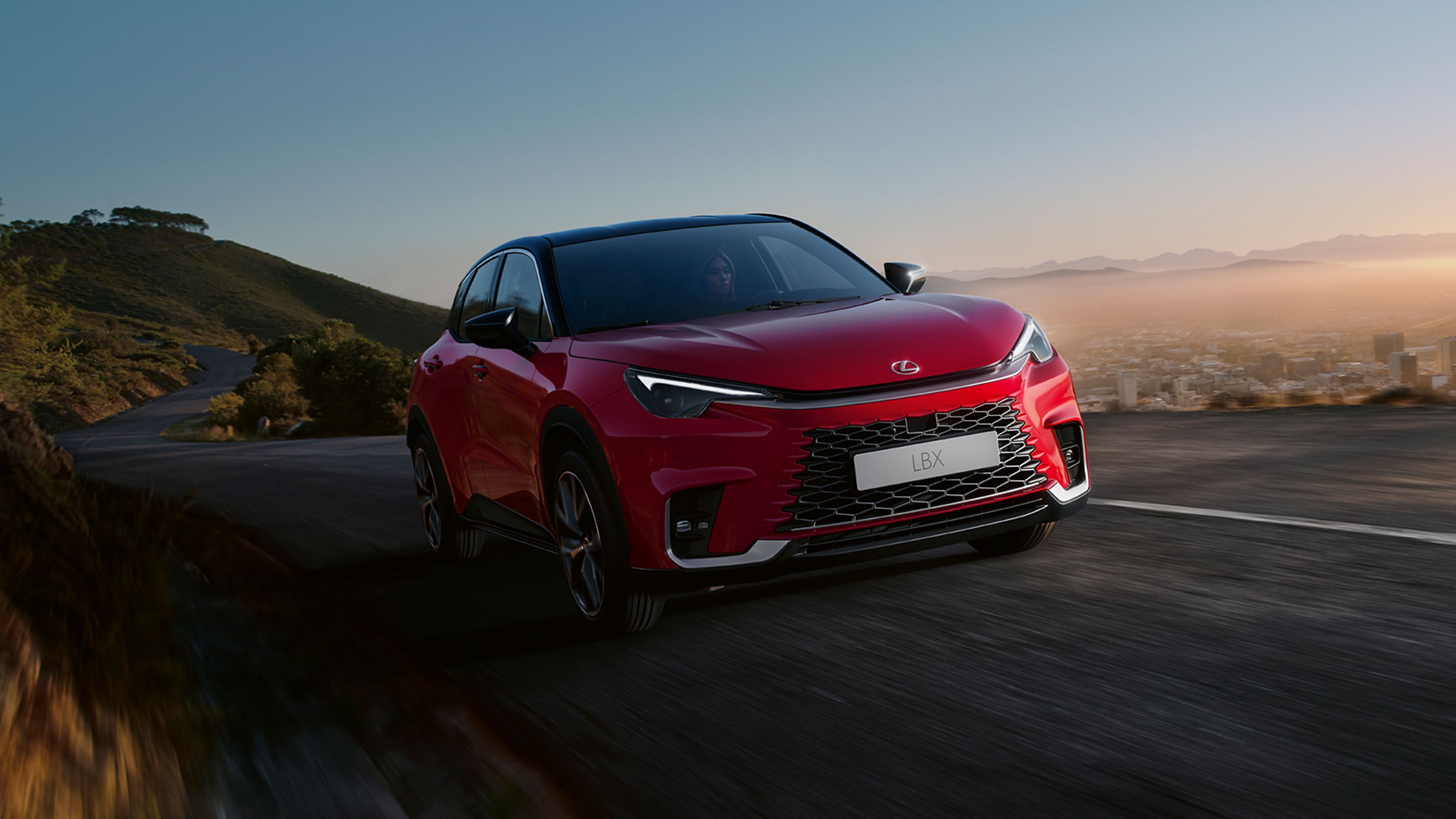 Ein roter Lexus LBX f&auml;hrt auf einer Bergstra&szlig;e oberhalb einer Gro&szlig;stadt entlang
