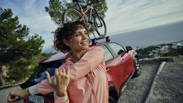 Eine Person steht vor einem Fahrzeug mit Fahrrad auf dem Dach und dehnt sich mit Lächeln im Gesicht.
