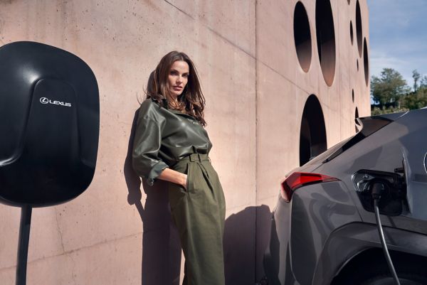 Eine Person lehnt lässig an einer Wand an der eine Lexus Wallbox befestigt ist. Neben ihr lädt ein geparkter Lexus.