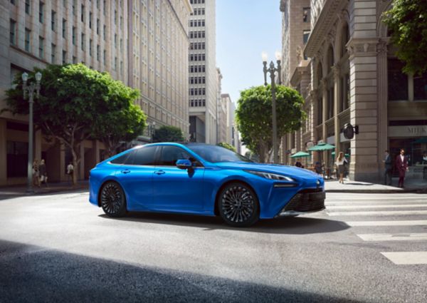 Ein blauer Toyota Mirai biegt an einer städtischen Kreuzung ab, im Hintergrund Fußgänger, ein Restaurant und Hochhäuser