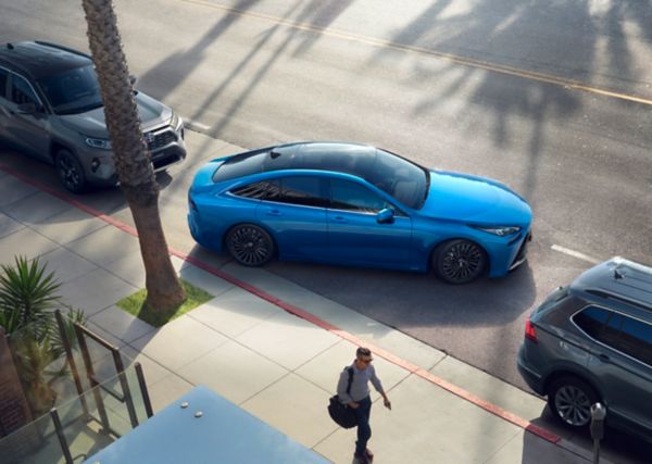 Ein blauer Mirai parkt seitlich zwischen zwei weiteren Autos an eine Straße ein. Daneben ist ein Baum und eine Person angeschnitten zu sehen.