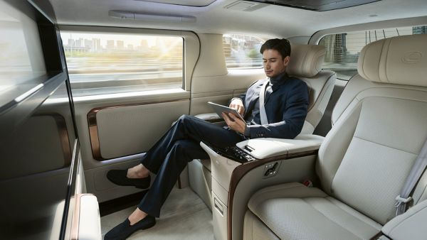 Ein Mann in einem maßgeschneiderten Anzug sitzt bequem in einem Lexus LM, konzentriert auf ein Tablet, mit Blick auf die Gebäude der Stadt durch das Fenster.