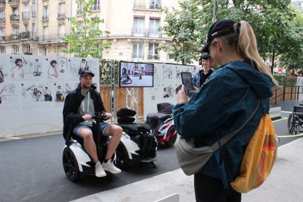 Ein Mann auf einer Art elektrischen Rollstuhl sitzend wird von einer Frau fotografiert, dahinter Informationsplakate und ein Bildschirm mit Erklärvideo