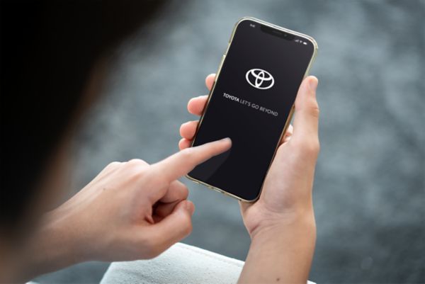A hand holds aloft a smartphone showing the My Garage page of the TOYOTA MyT app. Available Connected Services are shown.