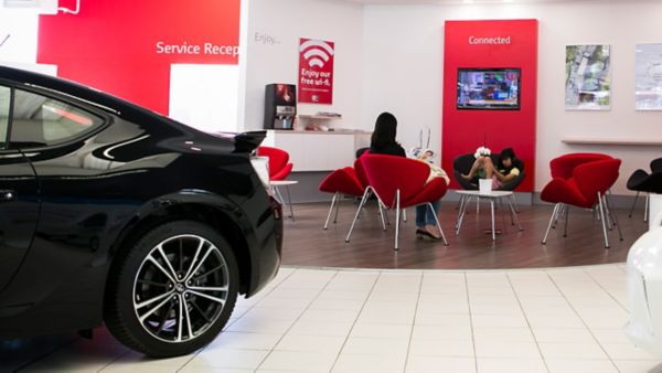 Ein modernes Auto im Vordergrund mit einem schlanken Design, während ein Wartebereich mit roten Stühlen und einem Fernseher ausgestattet ist, der Informationen in einem hellen, stilvollen Autohaus anzeigt.