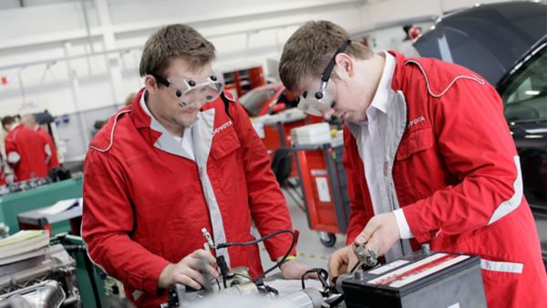 Zwei junge Techniker in roten Uniformen und mit Schutzbrillen arbeiten in einer Werkstatt gemeinsam an einem Automotor und konzentrieren sich dabei ganz auf ihre Aufgabe.