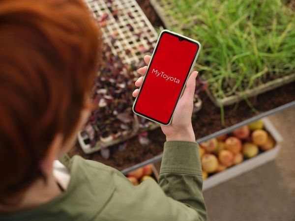Eine Person mit roten Haaren hält ein Smartphone mit der MyToyota-App auf einem leuchtend roten Hintergrund