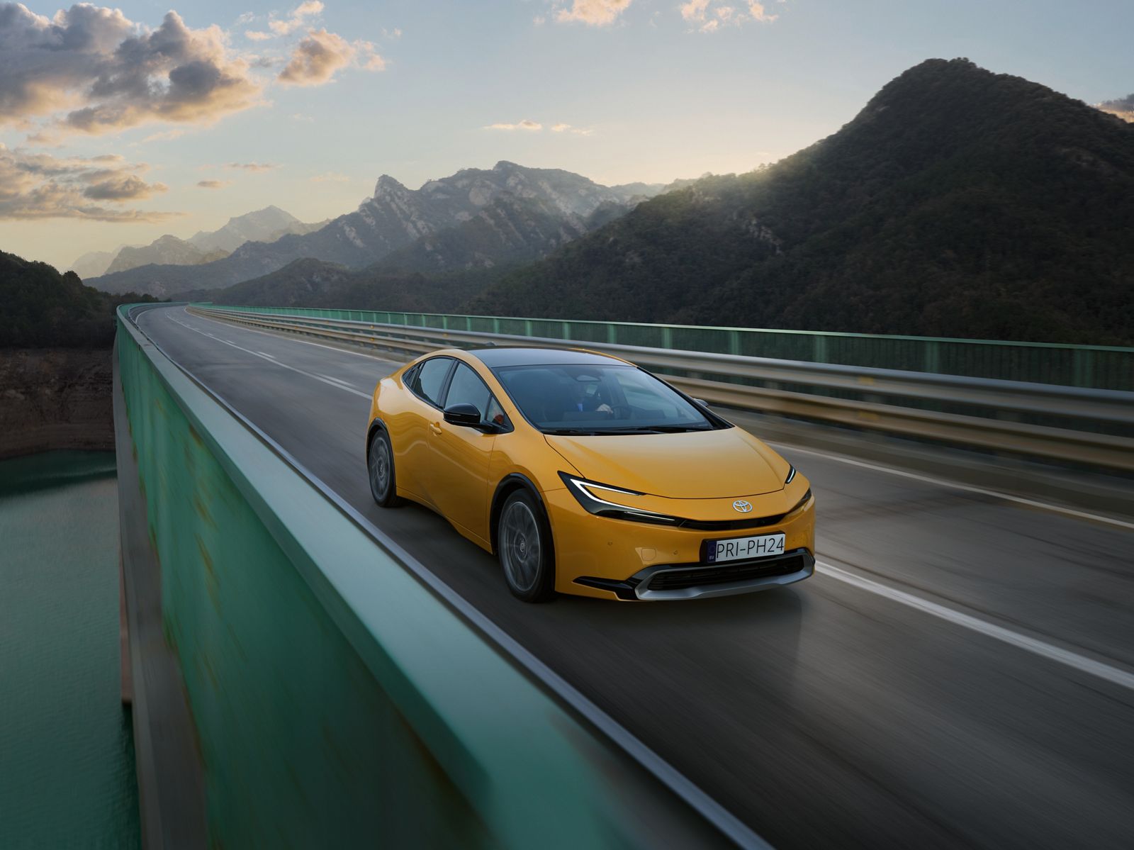 Gelber Prius fährt auf eine leeren Straße über eine Brücke, die Sonne steht tief hinter den Bergen im Hintergrund